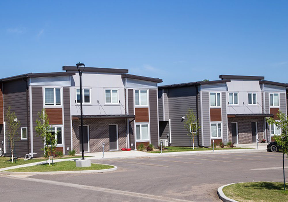 the-summit-seniors-apartment-brandon-smaller Exterior of The Groves townhouseson Tracey Street in Brandon, Manitoba. The two-storey townhouses are painted in dark earth tones and light grey, with brick facing around the entrances. Parking is available directly in front of each unit.